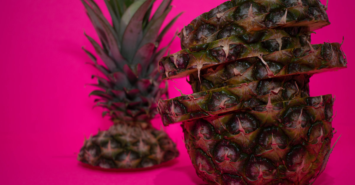 With a deep pink background, a whole pineapple sliced, with the top detached and placed to the left.