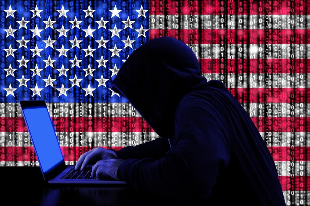 a hooded hacker using a lap top. A american flag with ones and zeros running through it can be seen in the background 