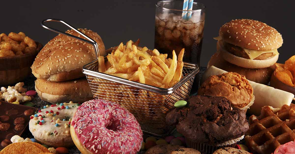 various unhealthy foods to depict a standard american diet. Foods in image include donuts, fries in a deep fryer basket, hamburgers and soda