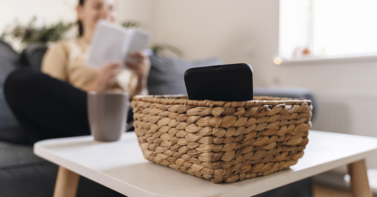 smartphone in a basket on a coffee table. Woman in background sitting on couch reading a book