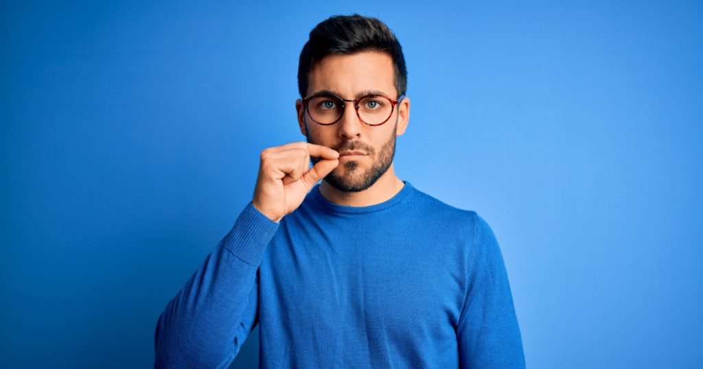man with beard wearing casual sweater and glasses over blue background mouth and lips shut as zip with fingers
