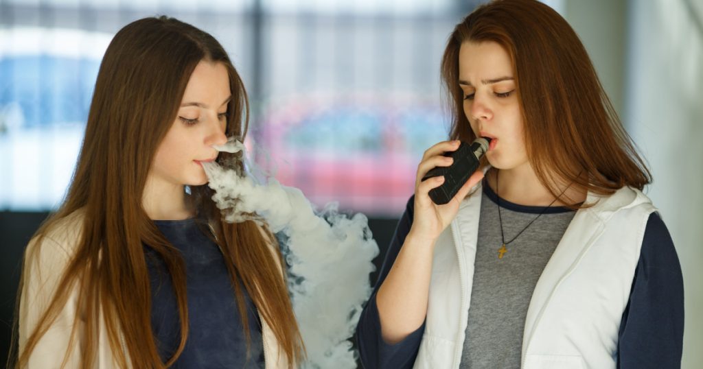 Vape teenagers. Two young cute girls in casual clothes smoke electronic cigarettes outdoors in the street in summer day. Bad habit that is harmful to health. Vaping activity.
