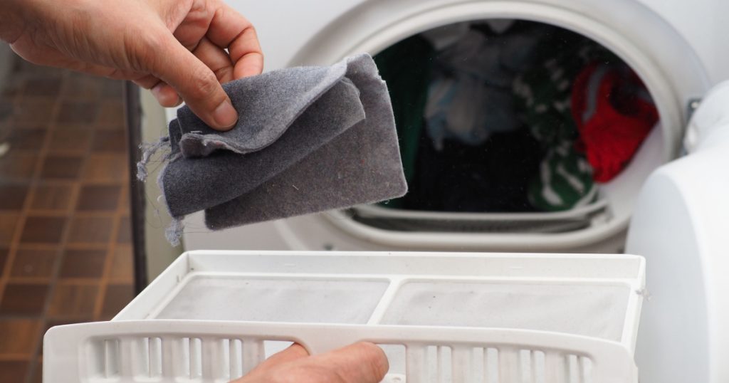 Woman’s hand removing lint from fluff filter of the tumble dryer on blurred background of clothes dryer with washed clothing. Laundry processes, Cleaning and care concept. (close up, selective focus)
