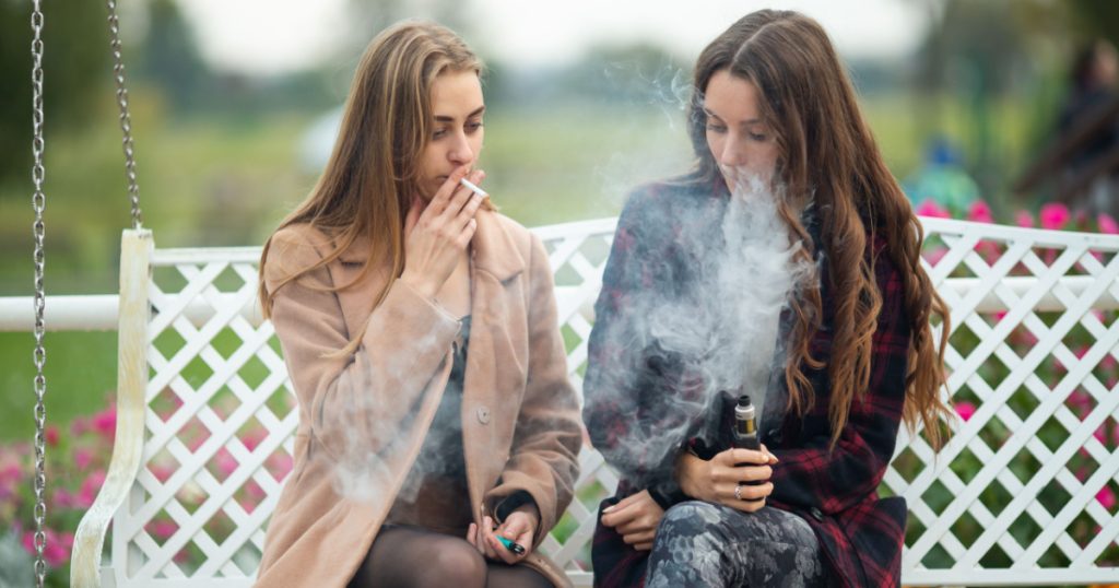 Smoking and vaping. Young beautiful white teeage girls in casual clothes sit on a vintage bench and smoke and vape cigarettes on the street in the evening. Electronic cigarette. Bad habit.
