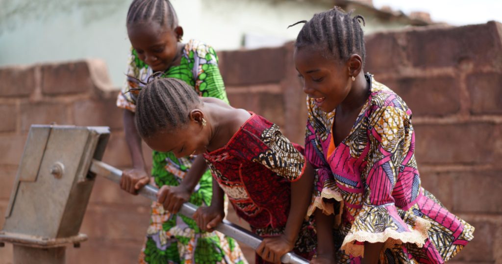 Three Smiling Little African Girls Busy Fetching Water At The Village Pump
