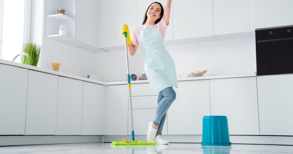 Full length photo of asian ethnicity housewife general cleaning disinfecting all surfaces infection safety hold mop clean floor wear gloves apron t-shirt jeans stand kitchen indoors
