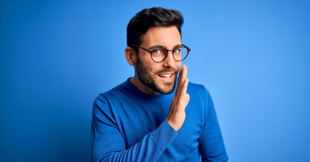 Young handsome man with beard wearing casual sweater and glasses over blue background hand on mouth telling secret rumor, whispering malicious talk conversation
