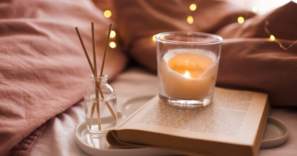 Burning candle with aroma sticks in bottle on tray with open book in bed over glowing Christmas lights close up. Cozy atmosphere at home. Good morning. Selective focus.
