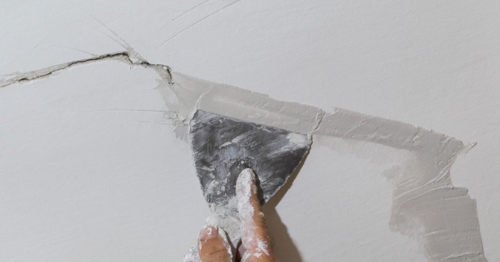 Worker fixing cracks on ceiling, spreading plaster using trowel
