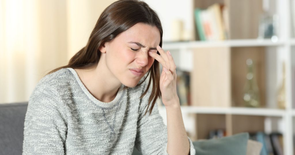 Woman scratching itchy eye sitting on a couch at home
