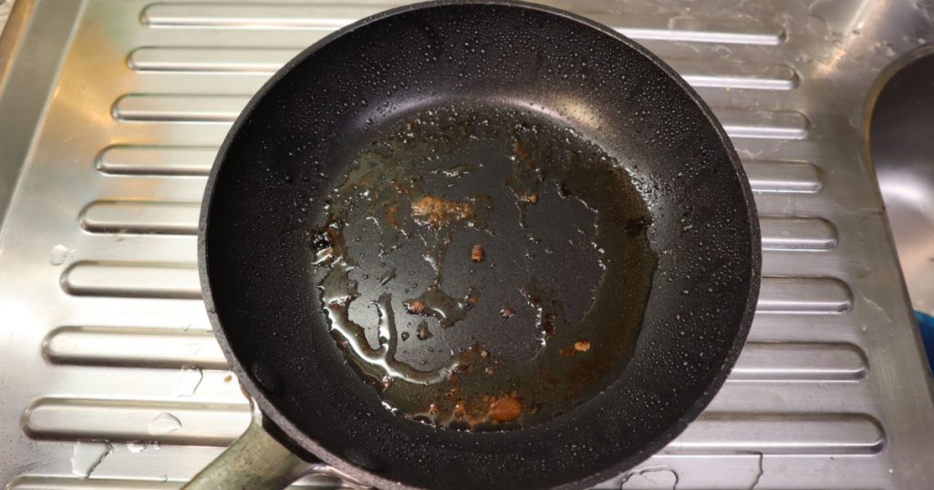A black, dirty non stick frying pan with grease splatter and food residue. A stainless steel drain can be seen in the background.
