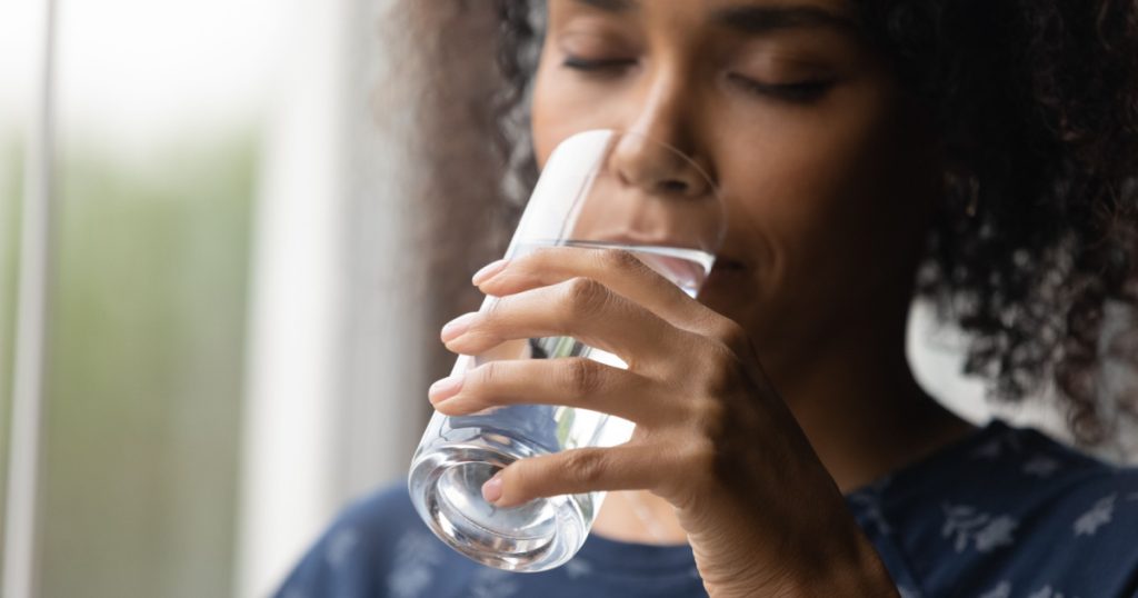 Closeup of young african woman drinking cool sparkling mineral water from glass hydrate having pleasure. Dehydrated black female feel thirsty drink swallow cold still aqua keep detox weight loss diet