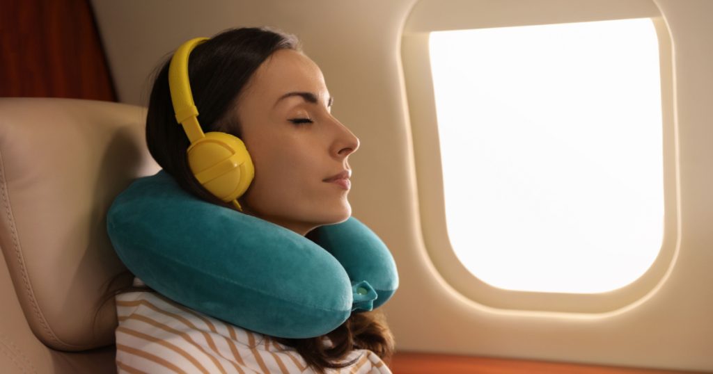 Young woman with travel pillow resting while listening to music in airplane during flight
