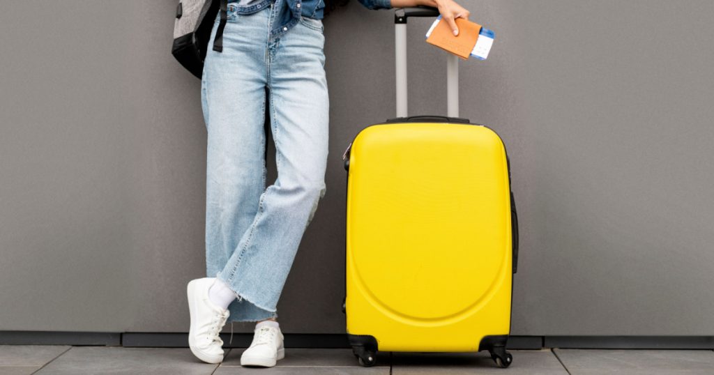 traveller standing over grey background, carrying yellow luggage and backpack, holding passport and flight tickets