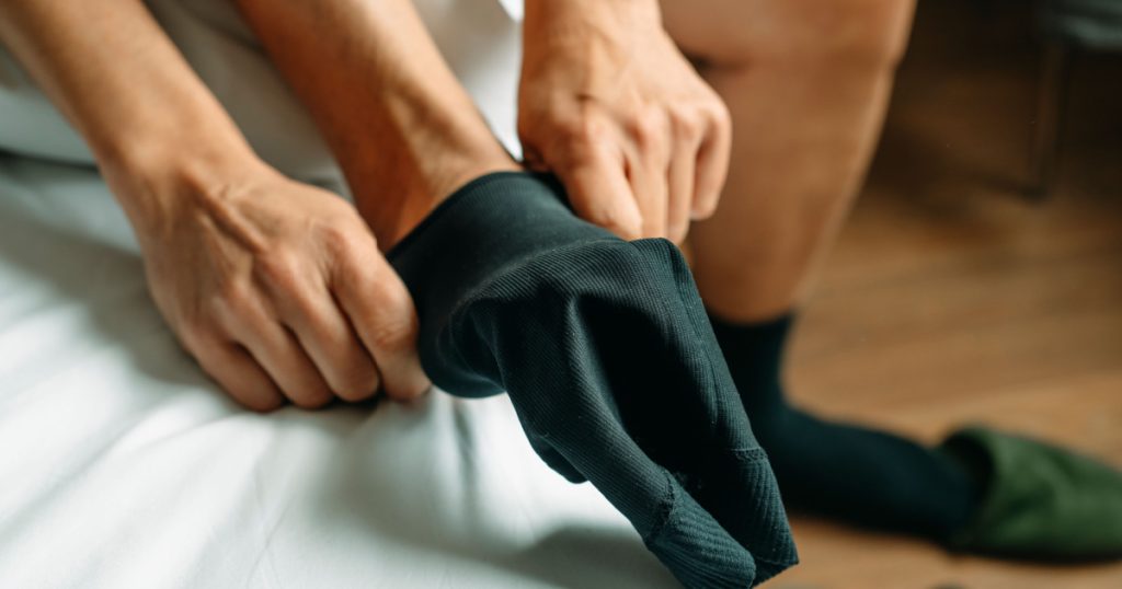 closeup of a man putting on a compression sock sitting on the edge of his bed at home
