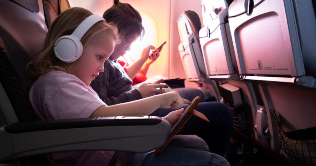 Family journey by plane. Little girl in wireless headphones using digital tablet, boy and woman entertaining with phones
