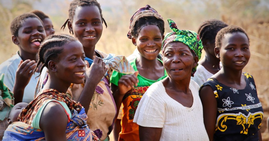 MALAWI, AFRICA A group of colorfully dressed happy healthy smiling African women of all ages gather at a clean water pump positive representation, cross generational mothers family child baby elderly
