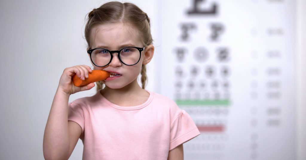 Happy cute girl in eyeglasses eating carrot, vitamin A for good vision, health
