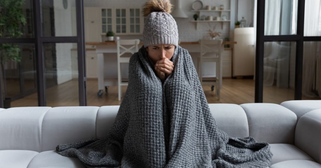  female sit on couch at freezing cooled studio flat in warm cap and blanket shiver tremble with cold