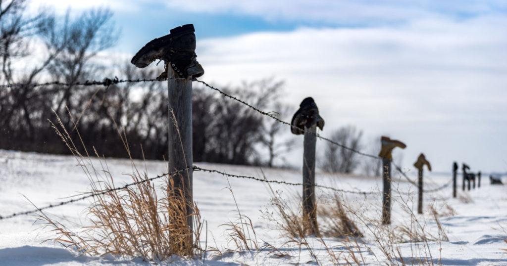 Old boots on top of a fence posts in a fence line south of Swift Current, Saskatchewan, Canada with tall grass poking out of the snow and blue sky
