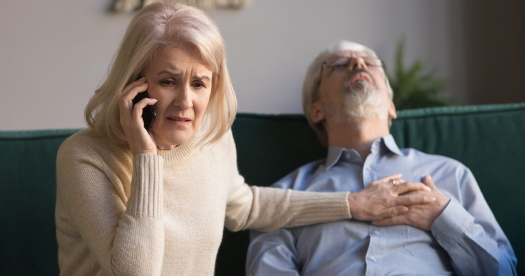 Desperate aged wife holds phone makes 911 emergency call while her sixty years husband lies on couch, man feeling crushing chest pain, loss of blood supply, shortness of breath, heart attack concept
