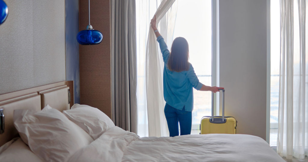 Traveler woman with suitcase standing near window and looking to a view from hotel room after check-in. Conceptual of travel and vacation holiday
