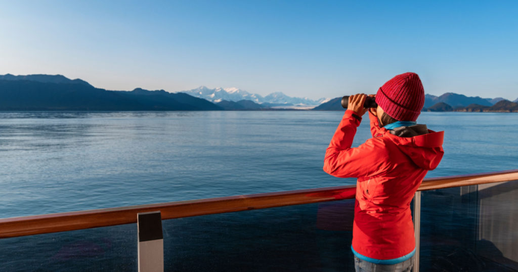 Vacation adventure. Alaska Glacier Bay cruise ship passenger looking at Alaskan mountains with binoculars exploring Glacier Bay National Park, USA. Woman on travel Inside Passage enjoying view
