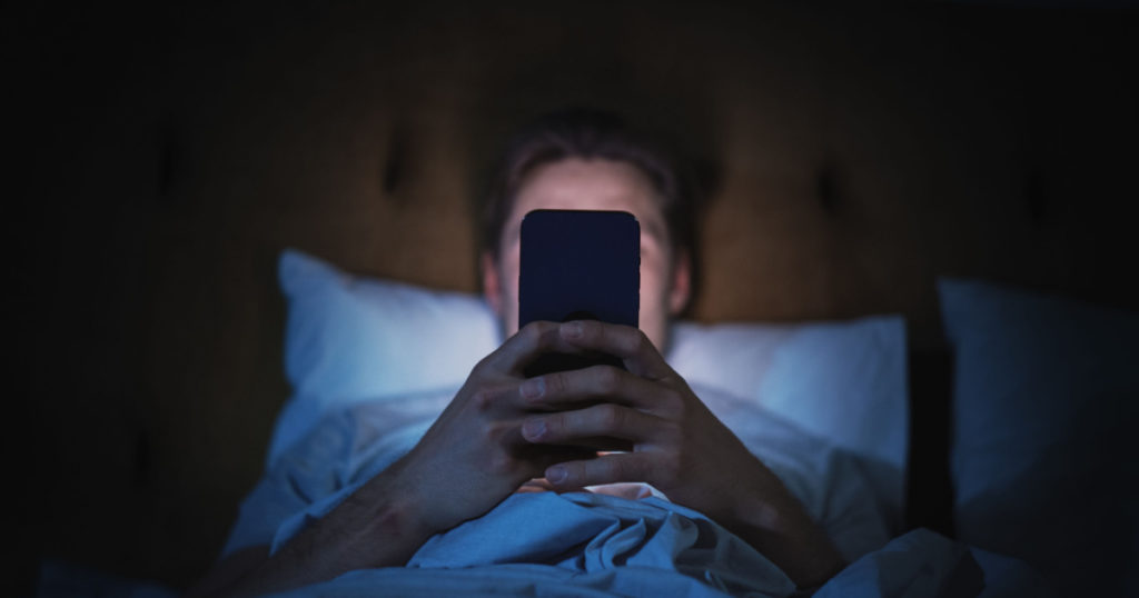 Man Uses Smartphone in Bed at Home at Night scrolling through social media 