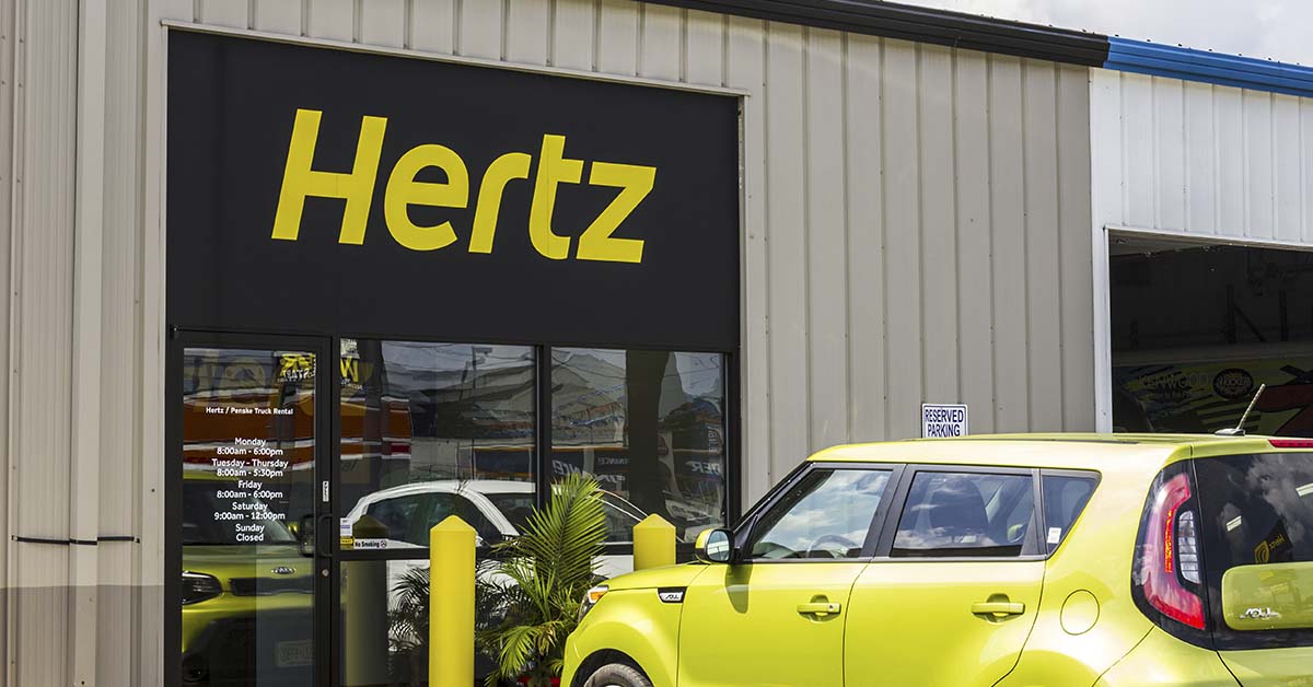 Outside of a Hertz car rental location. A Yellow Kia Soul is parked out front.