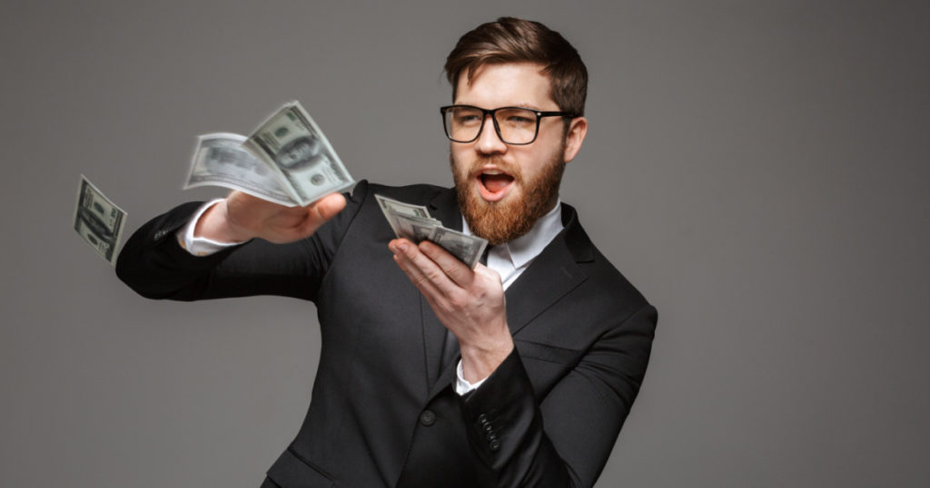 Portrait of a happy young businessman throwing out money banknotes isolated over gray background
