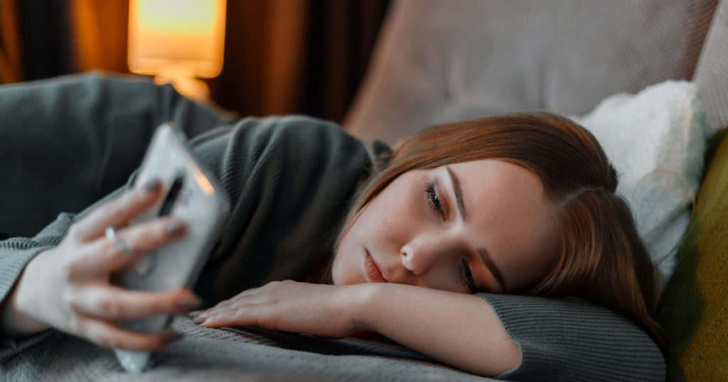 Sad teenage girl suffering from insomnia surfing Internet or chatting using smartphone at night lying on bed. Depression Internet addiction and depression in young woman. Long web banner.