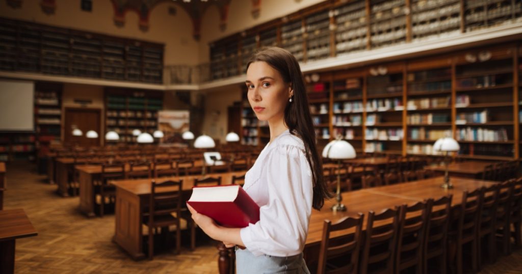 Portrait of attractive woman in white clothes with books in hands in beautiful public library posing at camera with serious face. Female student reads books in the student library.