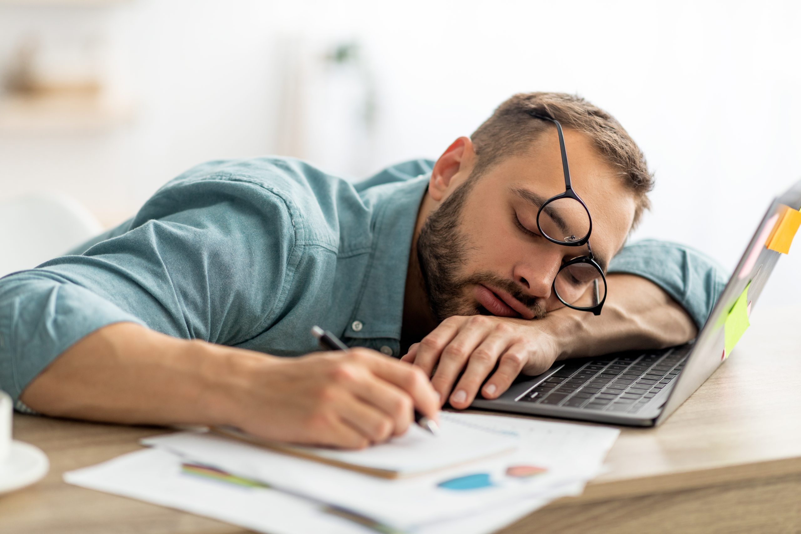 Exhausted millennial man sleeping on his office desk, next to laptop and documents, tired of overworking. Young male workaholic suffering from chronic fatigue at workplace
